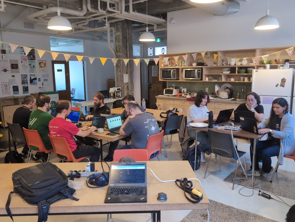 9 people sitting around 2 tables working at their respective laptops, with an unattended laptop in the foreground, taken from the opposite corner