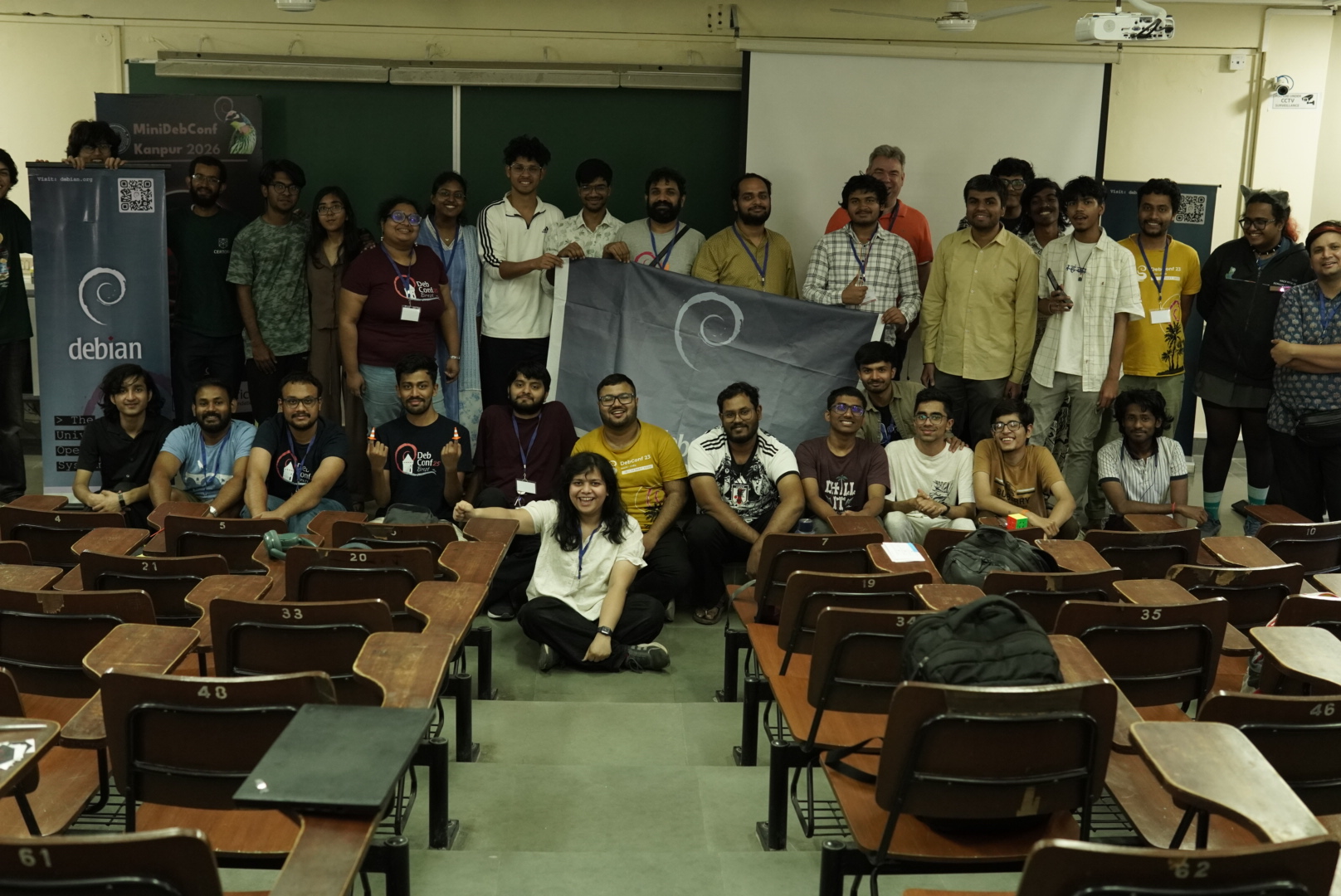 A classroom setting group photo. Multiple people looking at the camera. Some are standing, some are sitting. A poster Debian logo poster and standee can be seen. 
