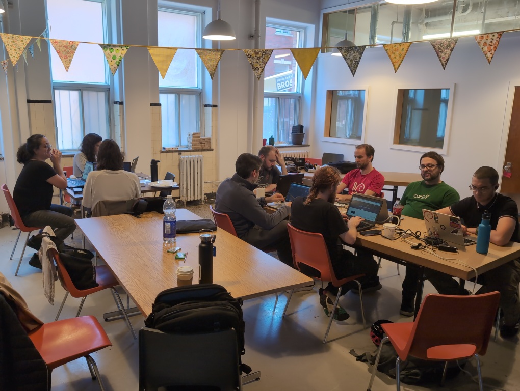 9 people sitting around 2 tables working at their respective laptops, taken from one corner