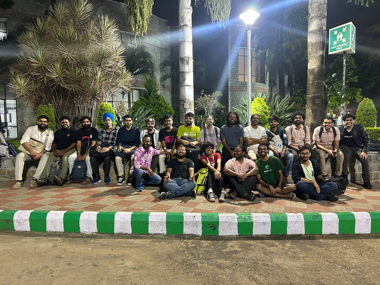 A group photo with one row of folks sitting on footpath and another behind then. Some trees can be seen in the backgroud. Everyone is smiling towards the camera.