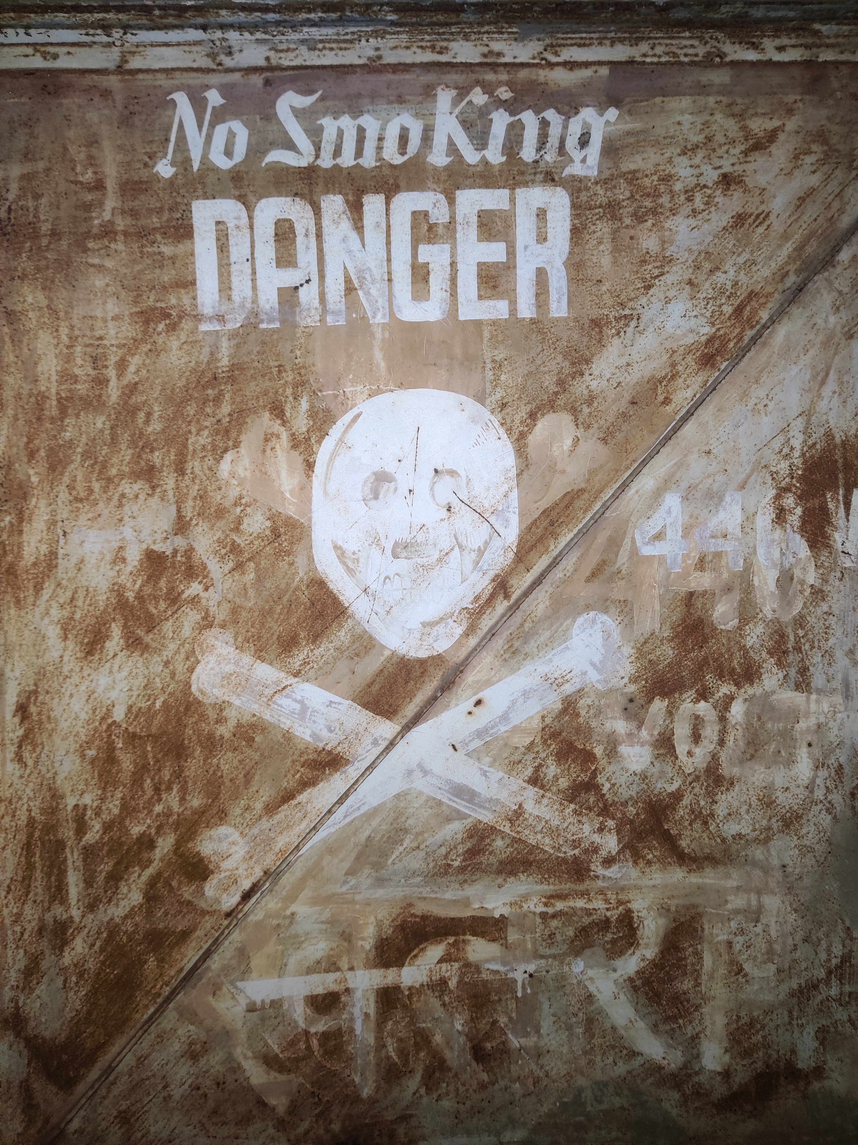 Picture depicting a rusted door with "No SmoKing" and "DANGER" written on it along with a skull and bones, likely leading to a power substation