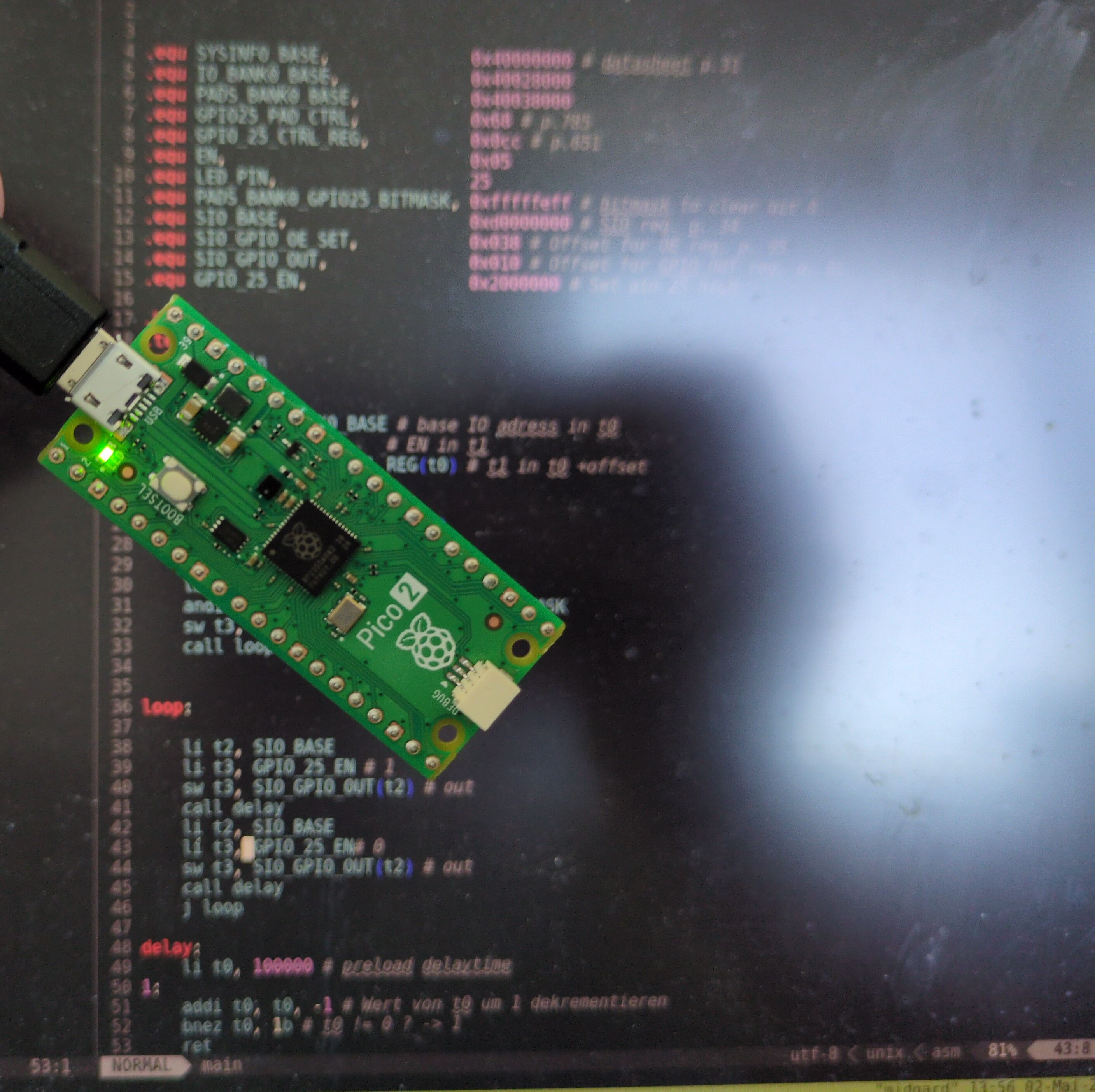 A Pico2 with its onboard LED on.  In the background a neovim window with riscv ASM code can be seen